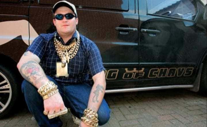 A screenshot of Michael Carroll wearing a lot of gold jewelry on his YouTube channel next to his large black car. 
