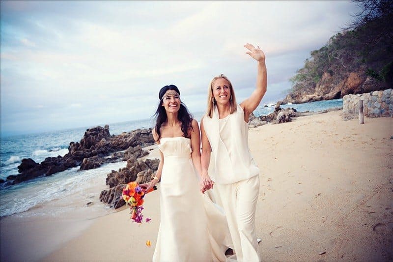 Jessica Clark and Lacey Stone in beautiful wedding outfits walking down the beach holding hands. 