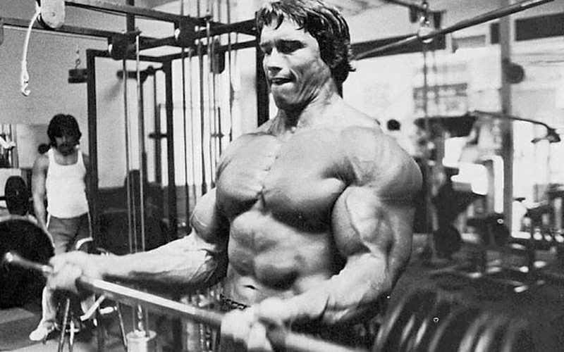A black and white photo of Arnold Schwarzenegger lifting at the gym. 