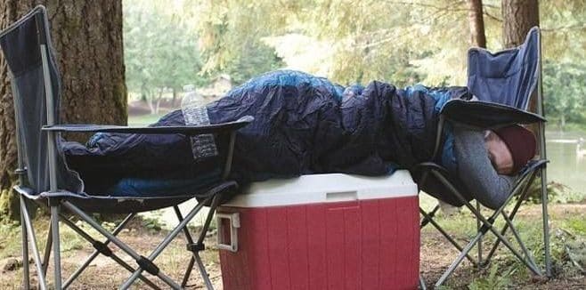 man lying on top of a cooler between two chairs outside