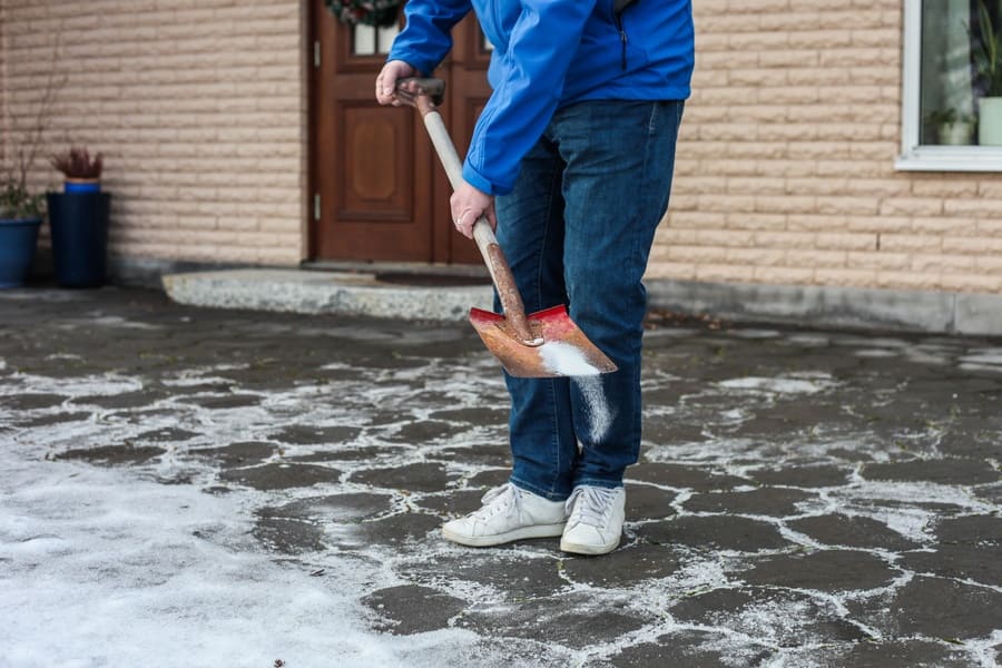 A man is using a shovel to sprinkle road salt on a stone walkway. 