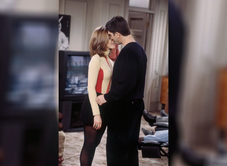Rachel and Ross standing in his living room with a short black shirt and a tan and red turtleneck shirt. 