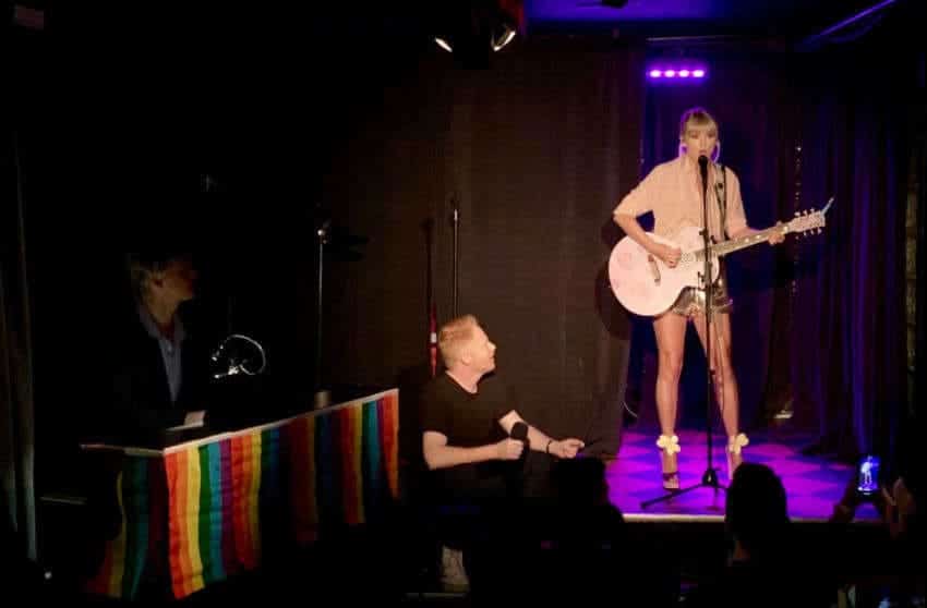 Taylor Swift is performing with Jesse Tyler Ferguson standing next to the stage holding a mic. 