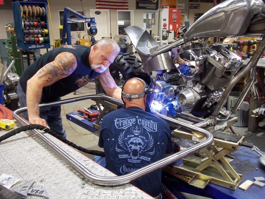 Paul Teutul Sr standing over someone who is working on a bike