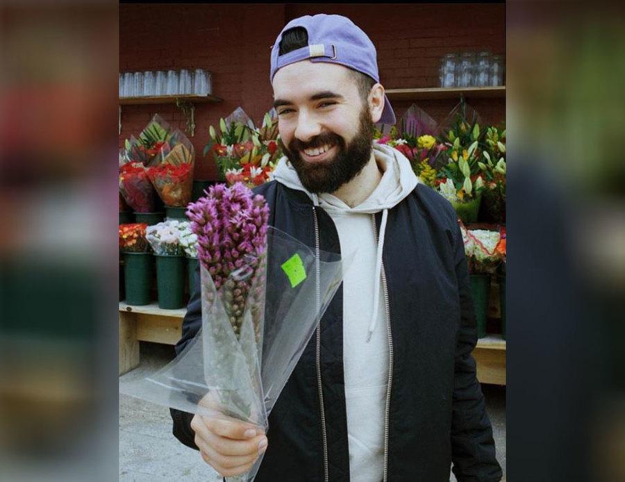 Jeremy is holding a bouquet of purple flowers outside of a flower shop. 