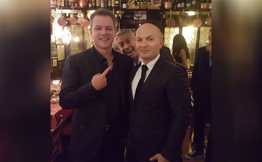 George Clooney behind Matt Damon and a waiter posing for a photograph 
