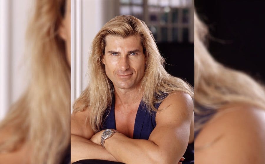 Fabio with his arms crossed wearing a blue tank top and his hair long and blonde