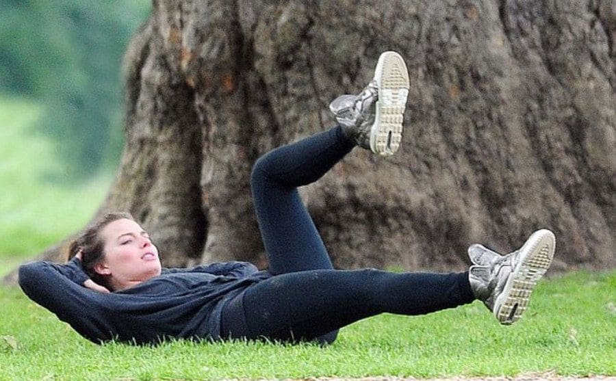Margot Robbie exercising in the grass near a large tree at the park 