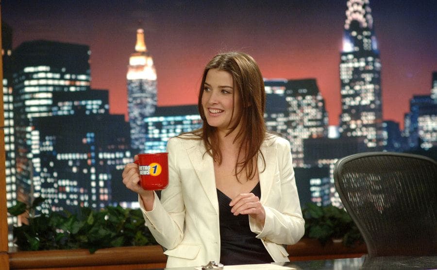 Cobie Smulders as Robin Scherbatsky sitting behind a news desk holding a cup of coffee