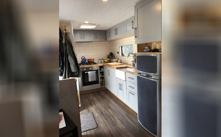 The mobile home kitchen with a grey and black fridge, light grey cabinets, white tile backwash, and light wood countertops 