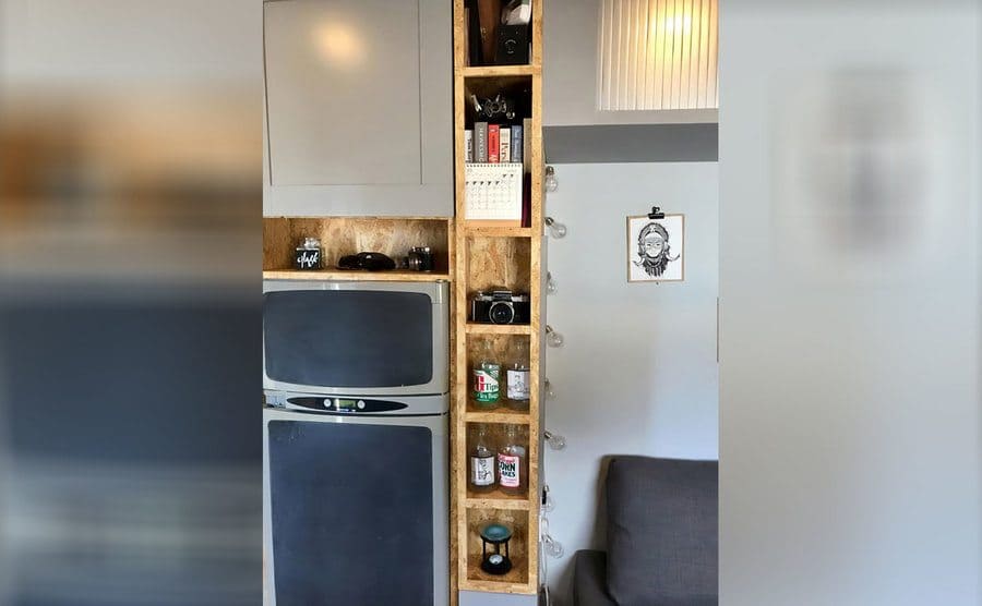 A photograph of the skinny shelf next to the fridge that goes from the floor to the ceiling with empty glass bottles, an old camera, and books sitting on the shelves