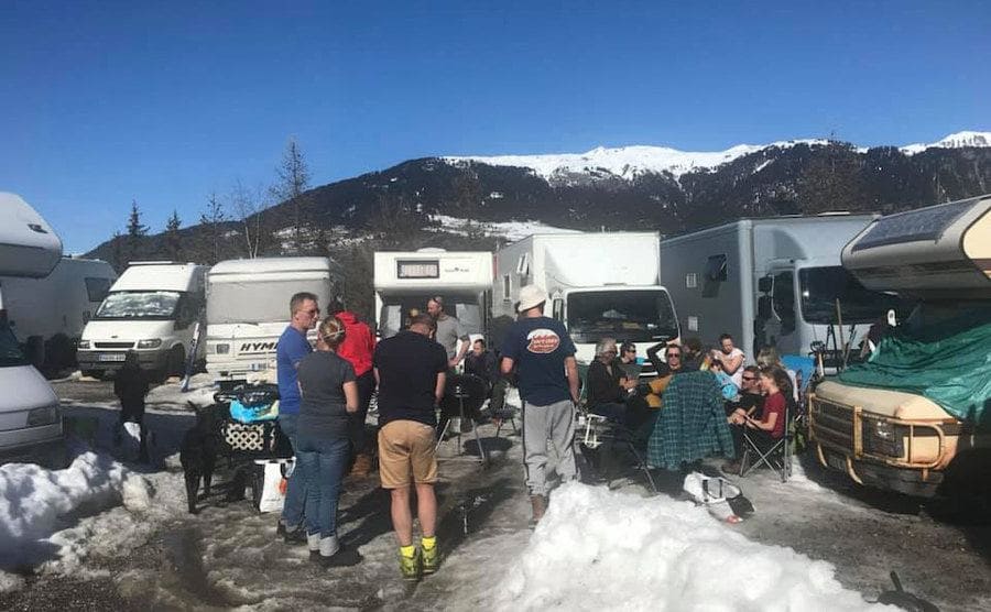 A snow-covered parking lot with RVs parked around a grill with everyone hanging around 