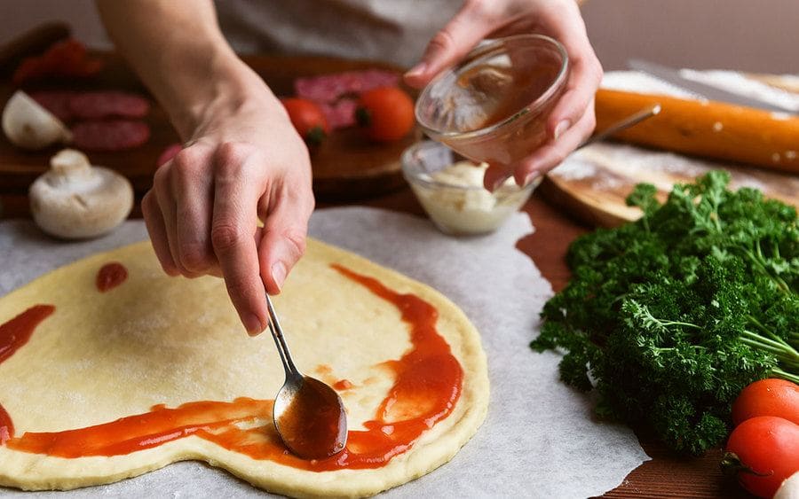 A woman spreading sauce on her homemade pizza dough in the shape of a heart