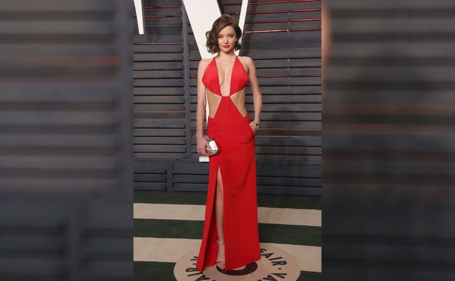 Miranda Kerr in a long red v-neck dress with the sides of her midriff exposed, a long slit, silver purse, and her hand in the pocket