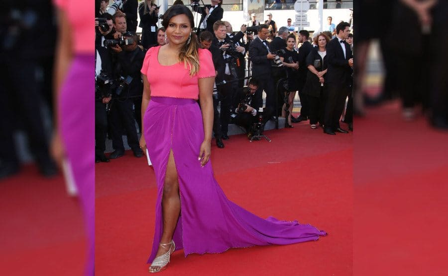 Mindy Kaling in a long dress with a peach-colored short-sleeved top and a purple bottom, a long slit, and silver strappy heels. 