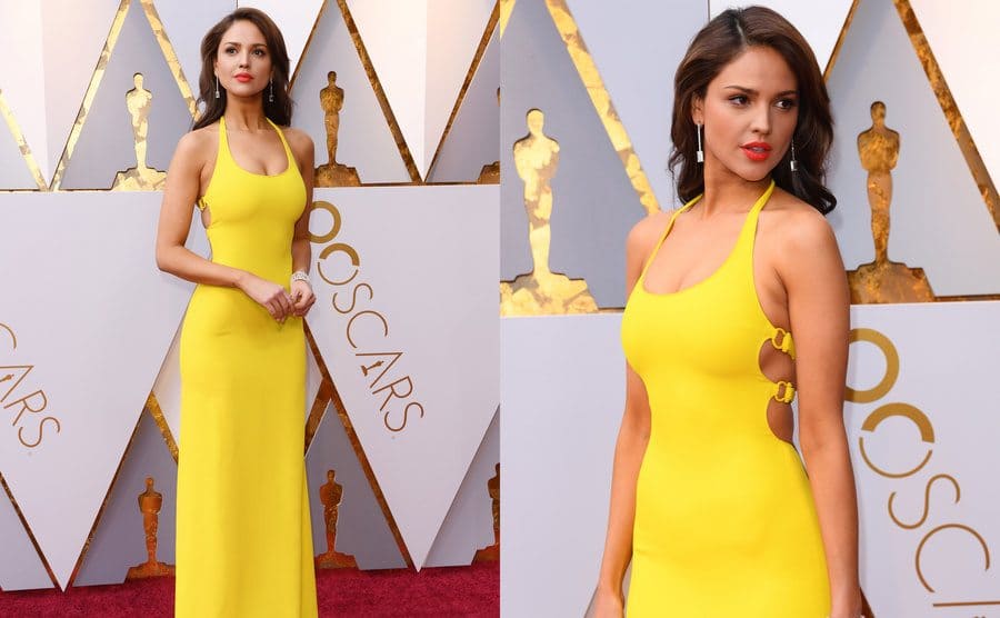 Eiza Gonzalez in a long yellow sleeveless dress with a boat neck. / Eiza Gonzalez closer up with the open side of her dress with a few straps going from front to back. 
