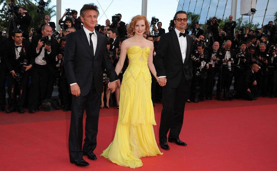 Jessica Chastain wearing a strapless yellow dress to the floor while holding Brad Pitt and Sean Penn's hands on the red carpet 