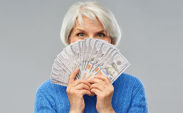 An older woman with grey hair holding a fan of $100 bills over her face 