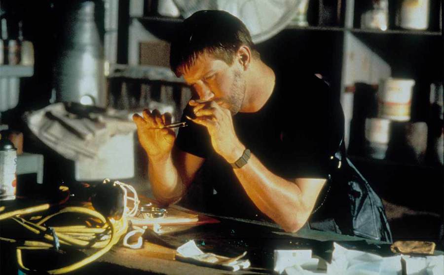 Stephen Baldwin sitting behind a desk looking at something small with a magnifying glass in the film The Usual Suspects