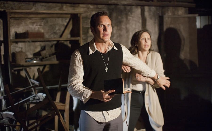 Patrick Wilson standing in front of Vera Farmiga with his arm out holding her back in a scene from The Conjuring 