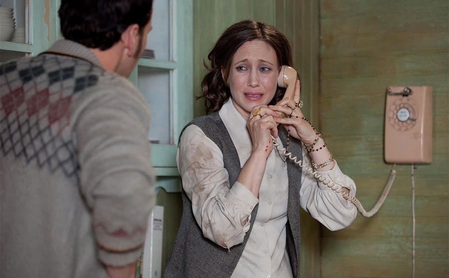 Vera Farmiga on the phone looking scared with Patrick Wilson standing with his back to the camera in a scene from The Conjuring 