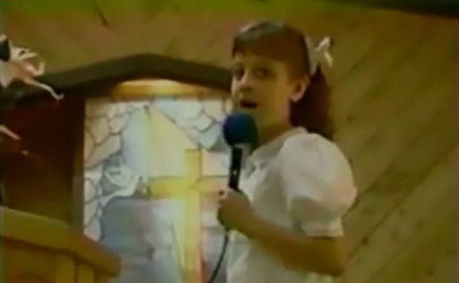 Beth as a child wearing a white dress holding a microphone and singing