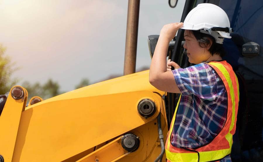 A woman wearing a white safety helmet standing in front of a backhoe