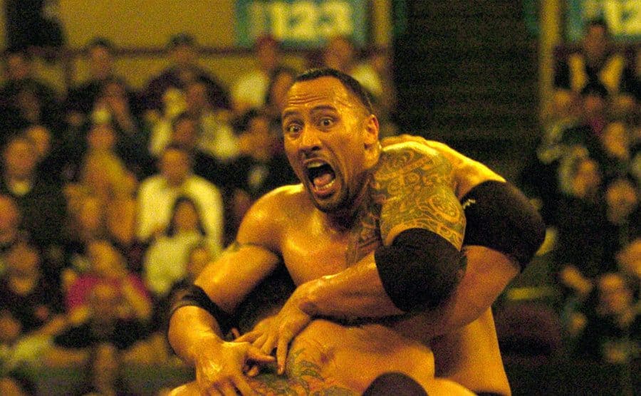 Dwayne Johnson being held by someone in a wrestling pose 