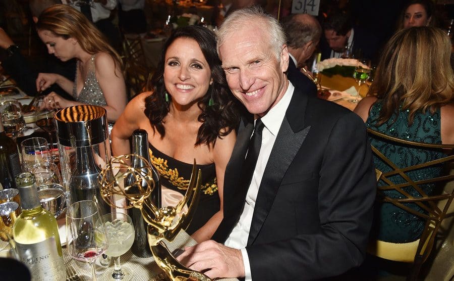 Julia Louis-Dreyfus and Brad Hall sitting at the dinner table at the Emmy Awards posing for a photo with her award sitting on the table 