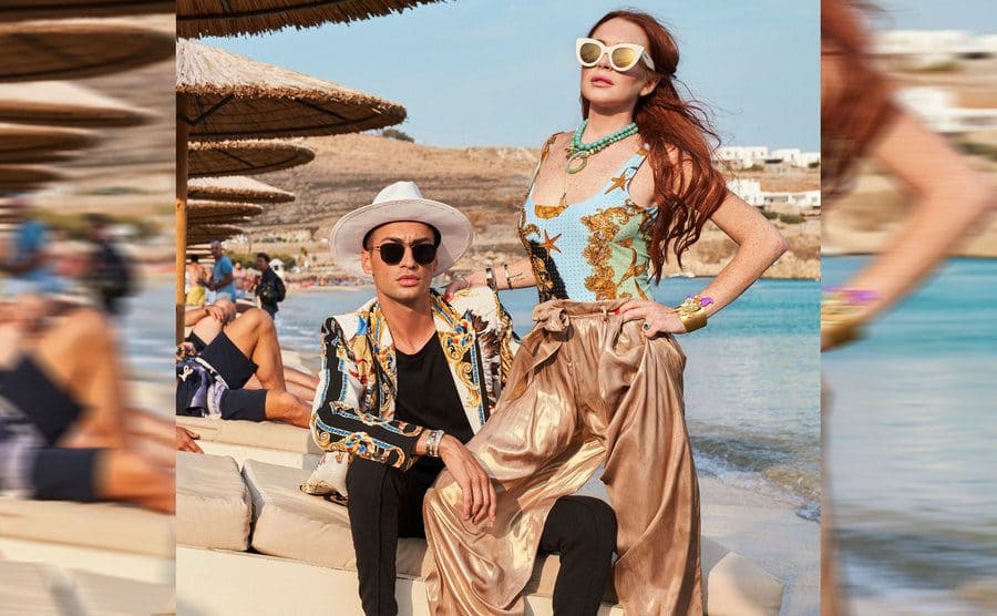 Lindsay Lohan posing standing next to Aristotle Polites sitting in a beach lounge chair in a promotional photograph from Lindsay Lohan’s Beach House 