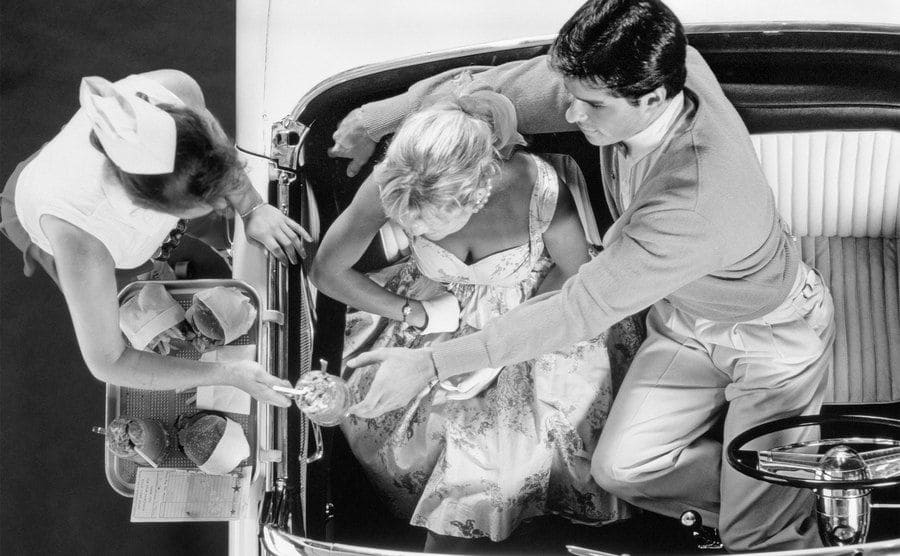 An aerial view of a couple in a convertible as part of a drive-in date having their food served to them in the car.