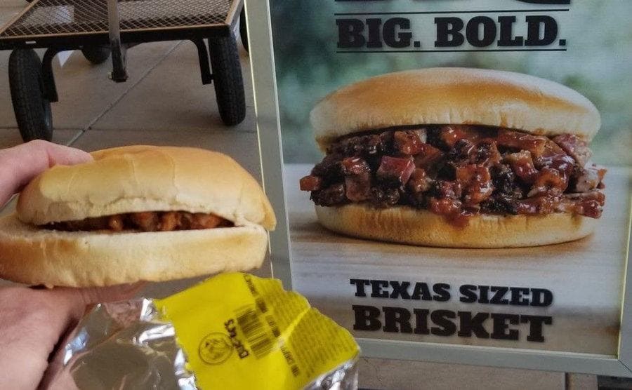 Enfoque de una triste hamburgesa delante de un cartel con una gran y cargada hamburgesa de carne