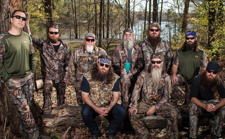 The cast of Duck Dynasty is sitting in the woods in camouflage attire. 