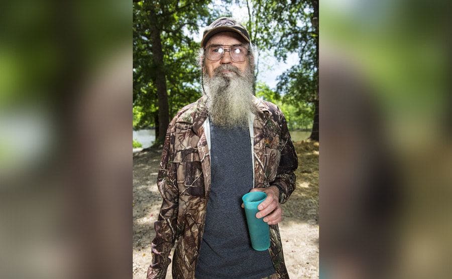 Uncle Si is holding his blue cup in the woods. 