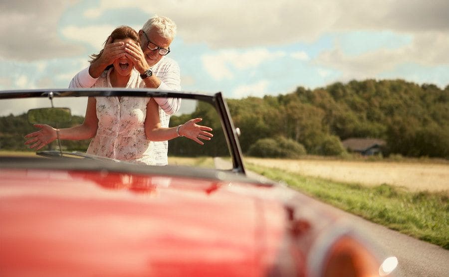 An older man is covering a woman’s eyes as he is about to give her a convertible.