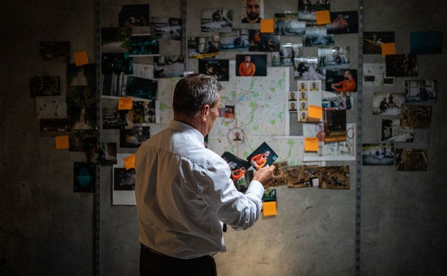 Rear view of gray-haired police detective looking pictures in front of the wall with a map, pictures, and adhesive notes on it, searching for a lead on his case.