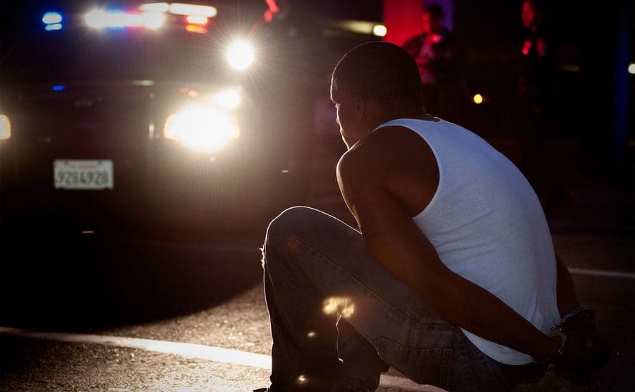 Police shining lights on handcuffed African man sitting on a curb.
