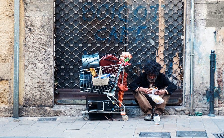 A beggar with his cart seated at the street eating a sandwich.