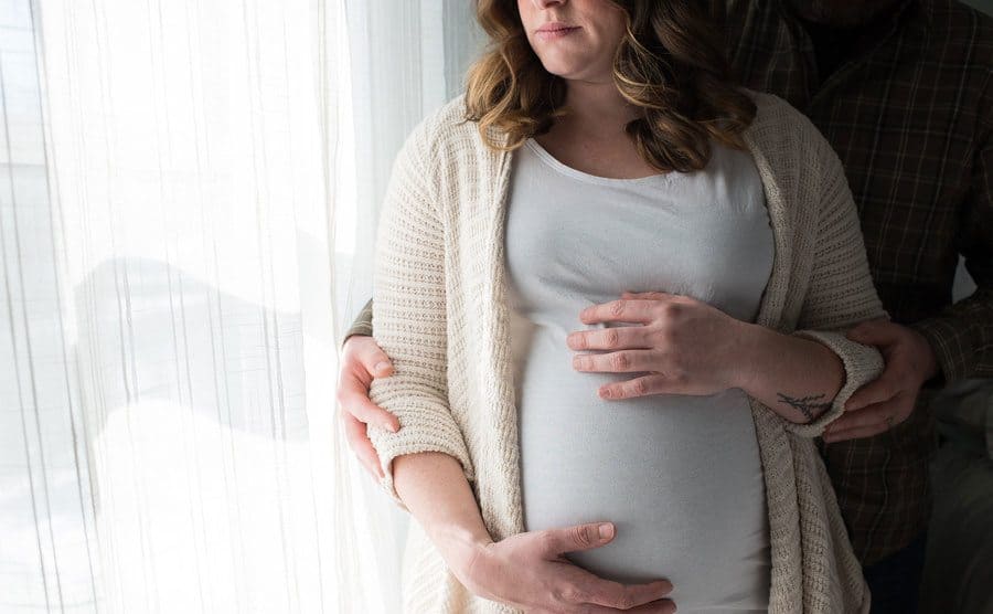 A pregnant woman stares out the window with her husband standing behind her. They both place their hands on the pregnant belly. 