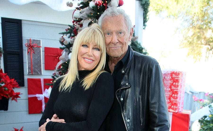 Suzanne Somers with her husband Alan Hamel posing in front of Christmas gifts on a porch 
