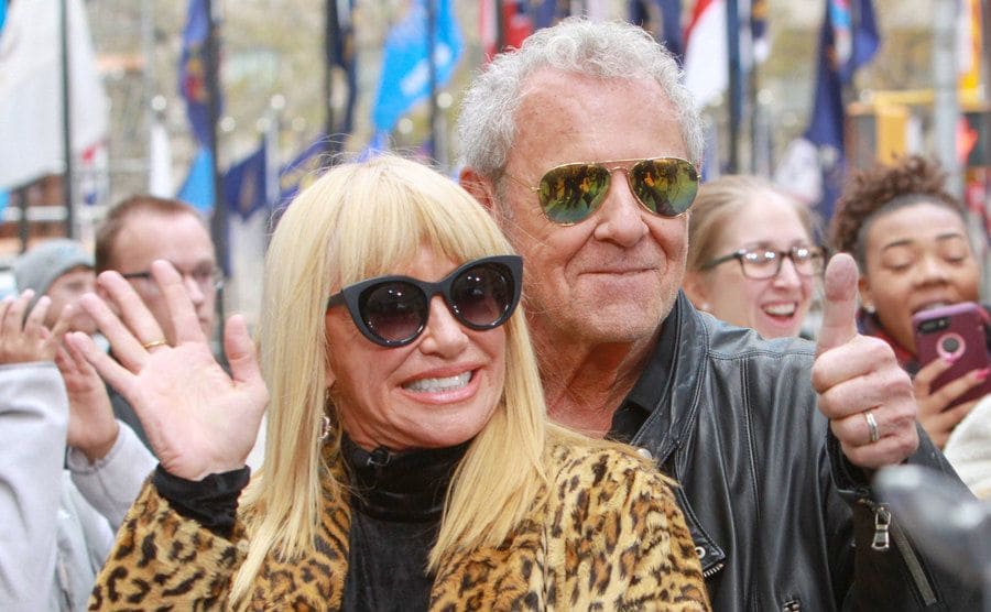 Alan Hamel and Suzanne Somers waving and giving a thumbs up outdoors in front of a crowd
