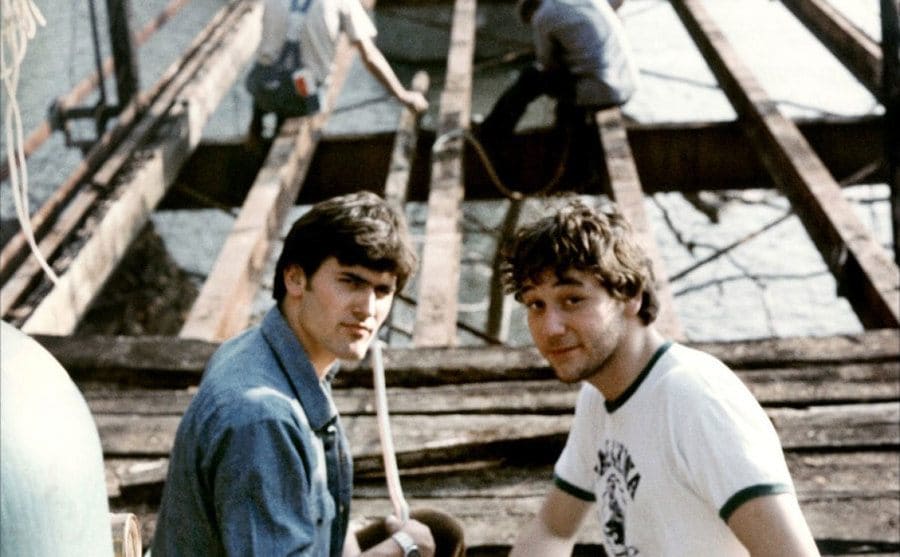 Bruce Campbell and Sam Raimi sitting on the side of a bridge which is being built 