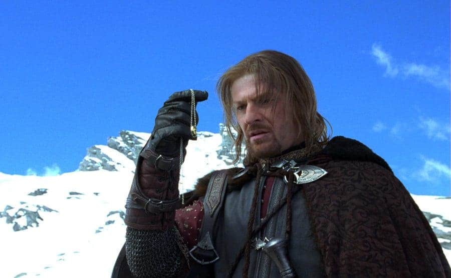 Sean Bean holding up a gold necklace in front of snow-covered mountains in a scene from Lord of the Rings 
