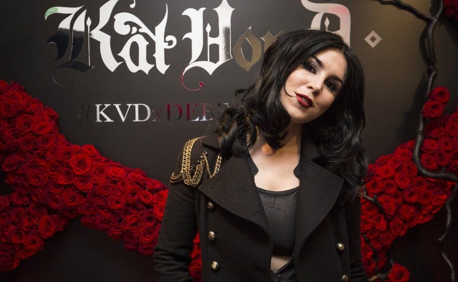 Kat Von D posing against a black wall with her beauty brand logo printed on it and roses all around 