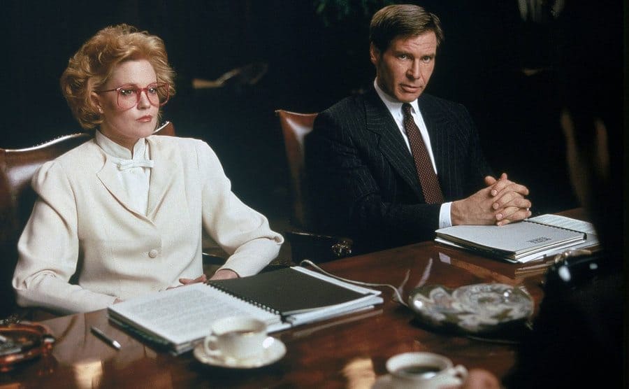 Melanie Griffith and Harrison Ford sitting behind a large wooden desk looking serious 