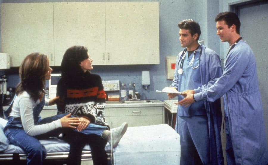 Jennifer Aniston y Courteney Cox sentadas en una cama de hospital con George Clooney y Noah Wyle como médicos 