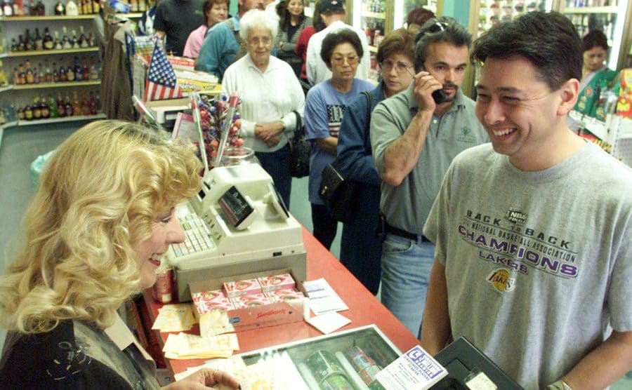 A woman is selling lotto tickets in the cashier as people line up at the store.