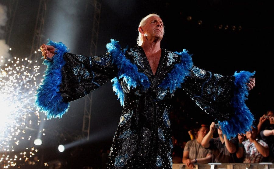 Ric Flair wearing a black leather and blue feathers glittery robe is being greeted by the crowd with flashing lights.