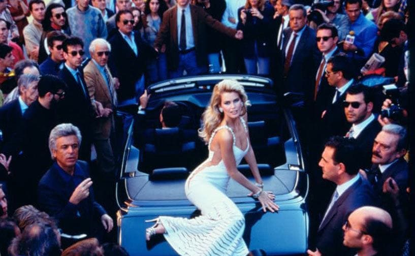 Claudia Schiffer poses on the truck of a car surrounded by a male crowd.