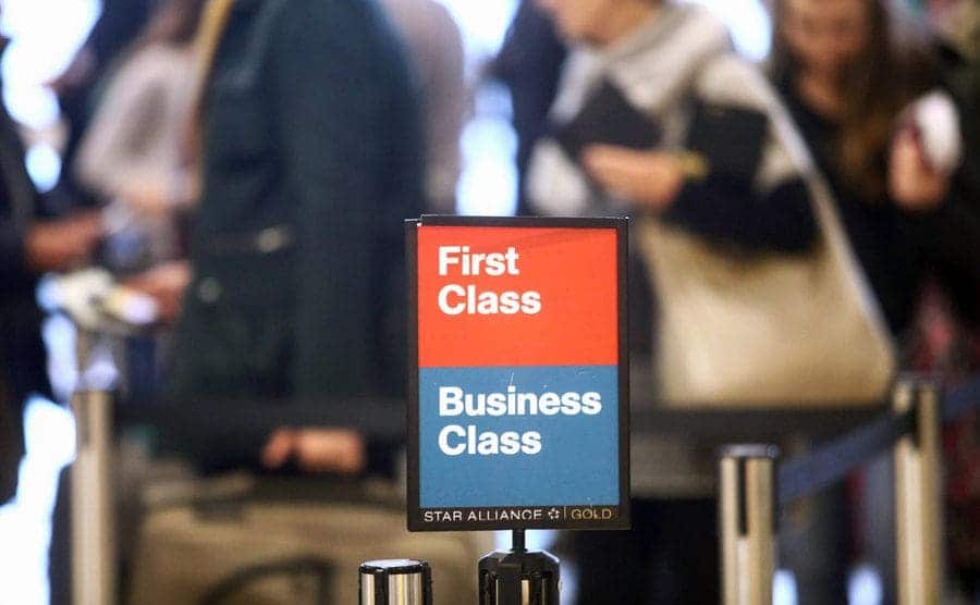 Passengers queue next to a ‘’First Class’’ and ‘’Business Class’’ sign at a departure gate inside.